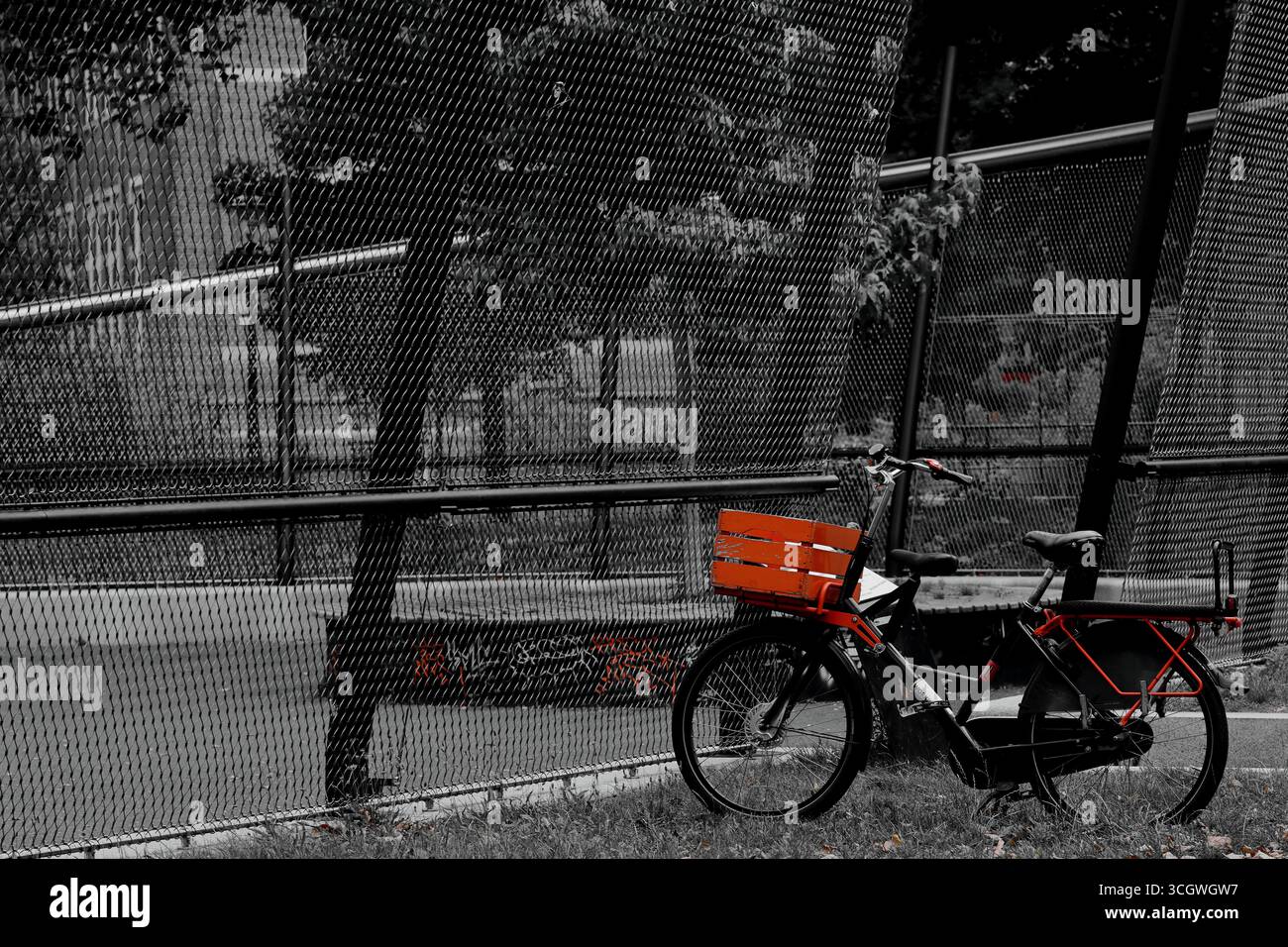 Vélo cargo orange-accent garé par une clôture à maillons de chaîne dans un parc de la ville, Amsterdam, pays-Bas, avec fond en couleur sélective monochrome urbaine. Banque D'Images