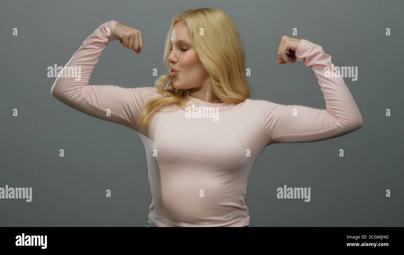 Femme fléchissant les muscles en toute confiance sur un fond gris lisse, portant une chemise rose décontractée, incarnant la force, l'autonomisation, la forme physique et la positiv Banque D'Images
