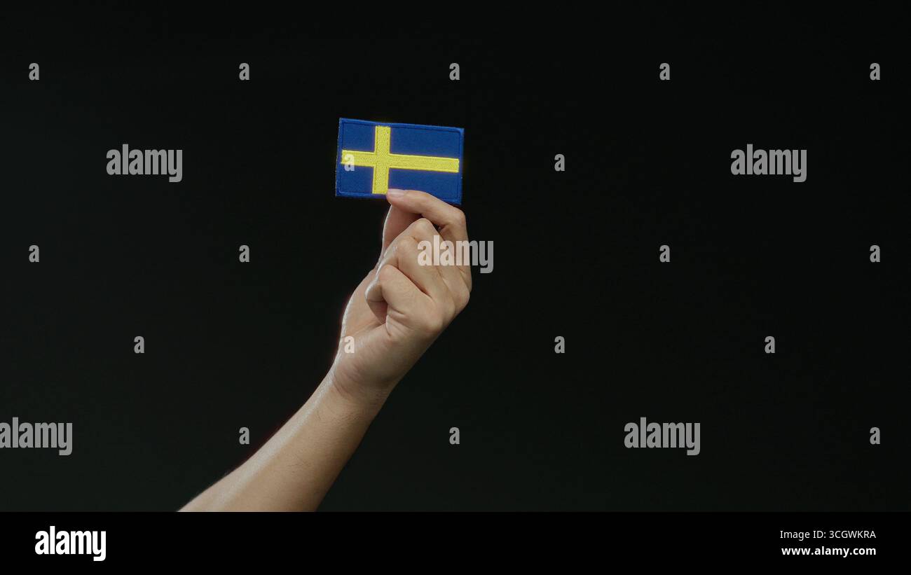 Main tenant un patch de drapeau suédois sur un fond noir, symbolisant la nationalité suédoise et la fierté, isolé et important dans la scène, capturin Banque D'Images