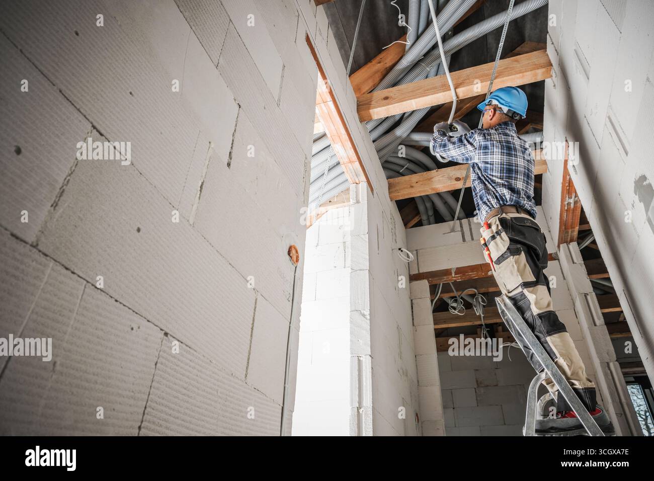 Un ouvrier de la construction utilise une échelle pour installer le câblage électrique dans le plafond d'un nouveau bâtiment. L'espace de travail présente des poutres apparentes et des matériaux Banque D'Images