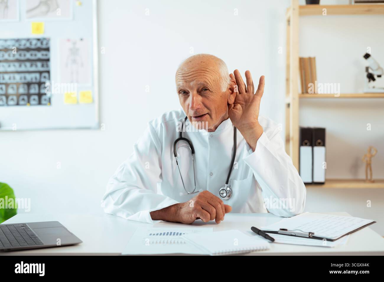 médecin mûr avec stéthoscope fonctionne sur un ordinateur portable regardant la caméra essayez d'entendre l'écoute de surentendre intentionnellement. Thérapeute senior faisant un appel vidéo, practi Banque D'Images