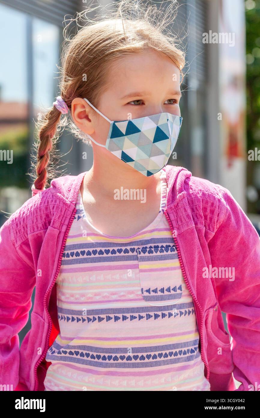 Fille d'âge scolaire portant un masque facial de protection de style moderne à l'extérieur le jour ensoleillé, veste rose élégante, cheveux tressés, précautions de sécurité contre les virus pandémiques Banque D'Images