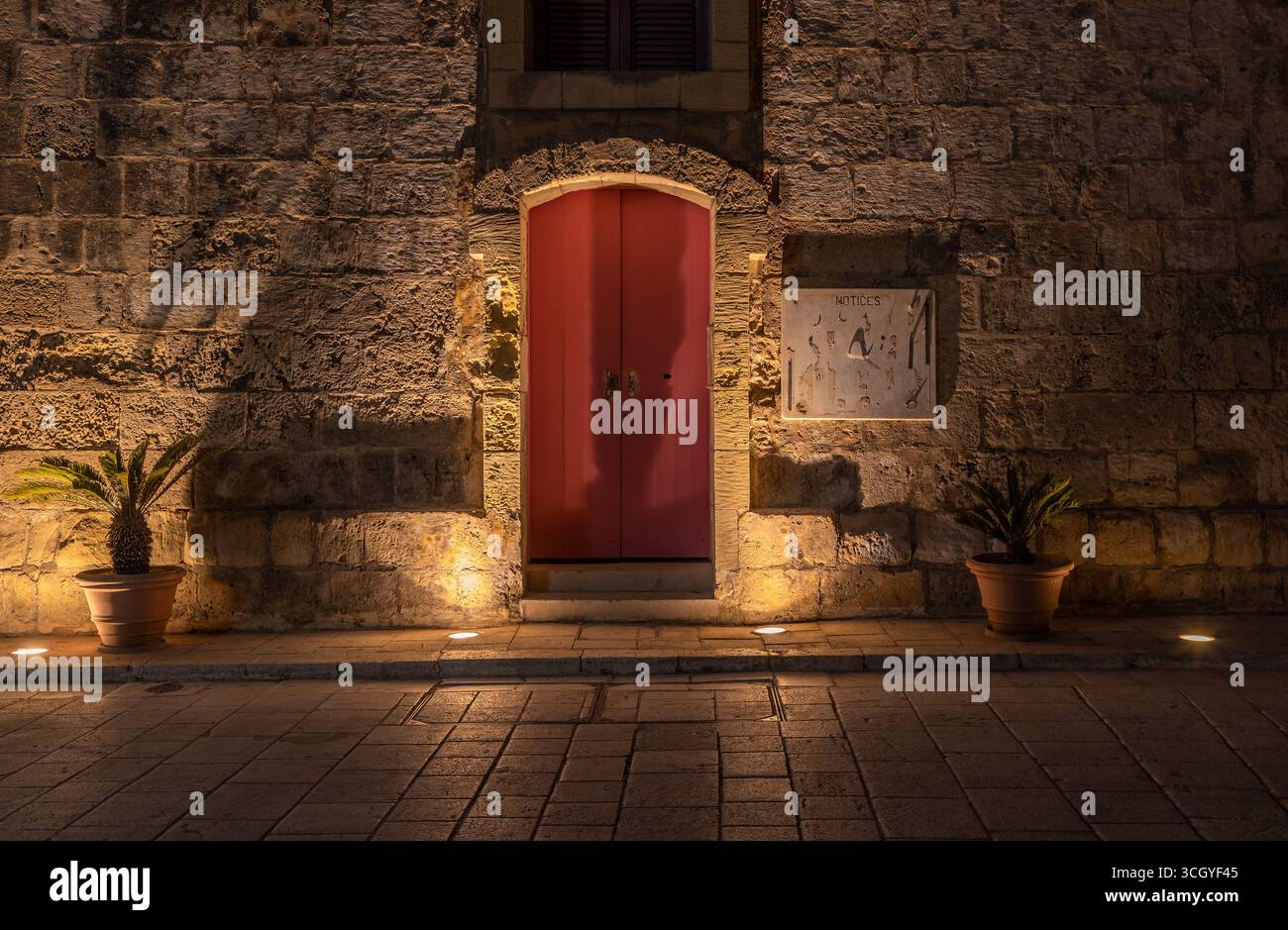 Porte rouge lumineuse située dans un mur de pierre historique avec des plantes en pot à Mdina, Malte. Banque D'Images