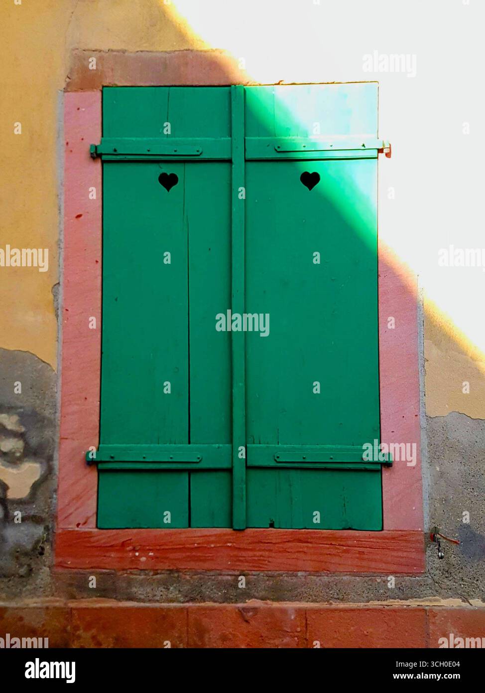 Fenêtre avec volets verts et détail coeur sur un mur de maison rouge corail et jaune avec des pierres naturelles d'Alsace, France Banque D'Images