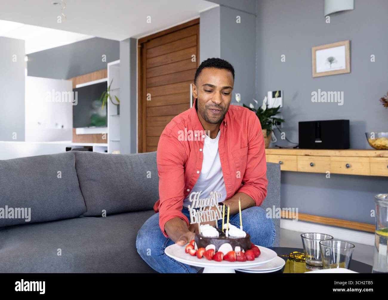 Homme afro-américain tenant un gâteau d'anniversaire avec des bougies allumées sur le canapé dans le salon, souriant Banque D'Images