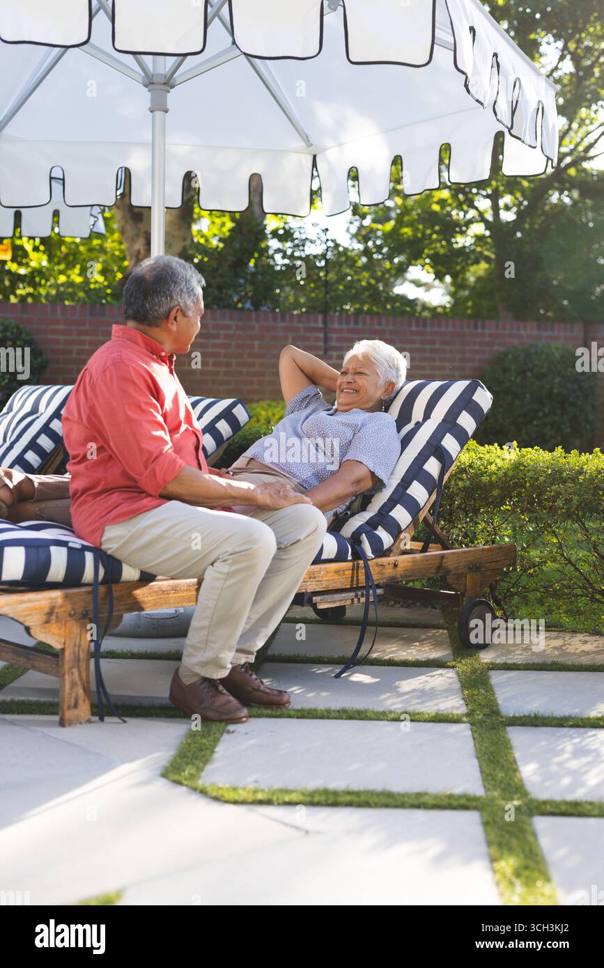 Vieux couple diversifié se relaxant sur des chaises longues sous parasol dans le jardin ensoleillé Banque D'Images