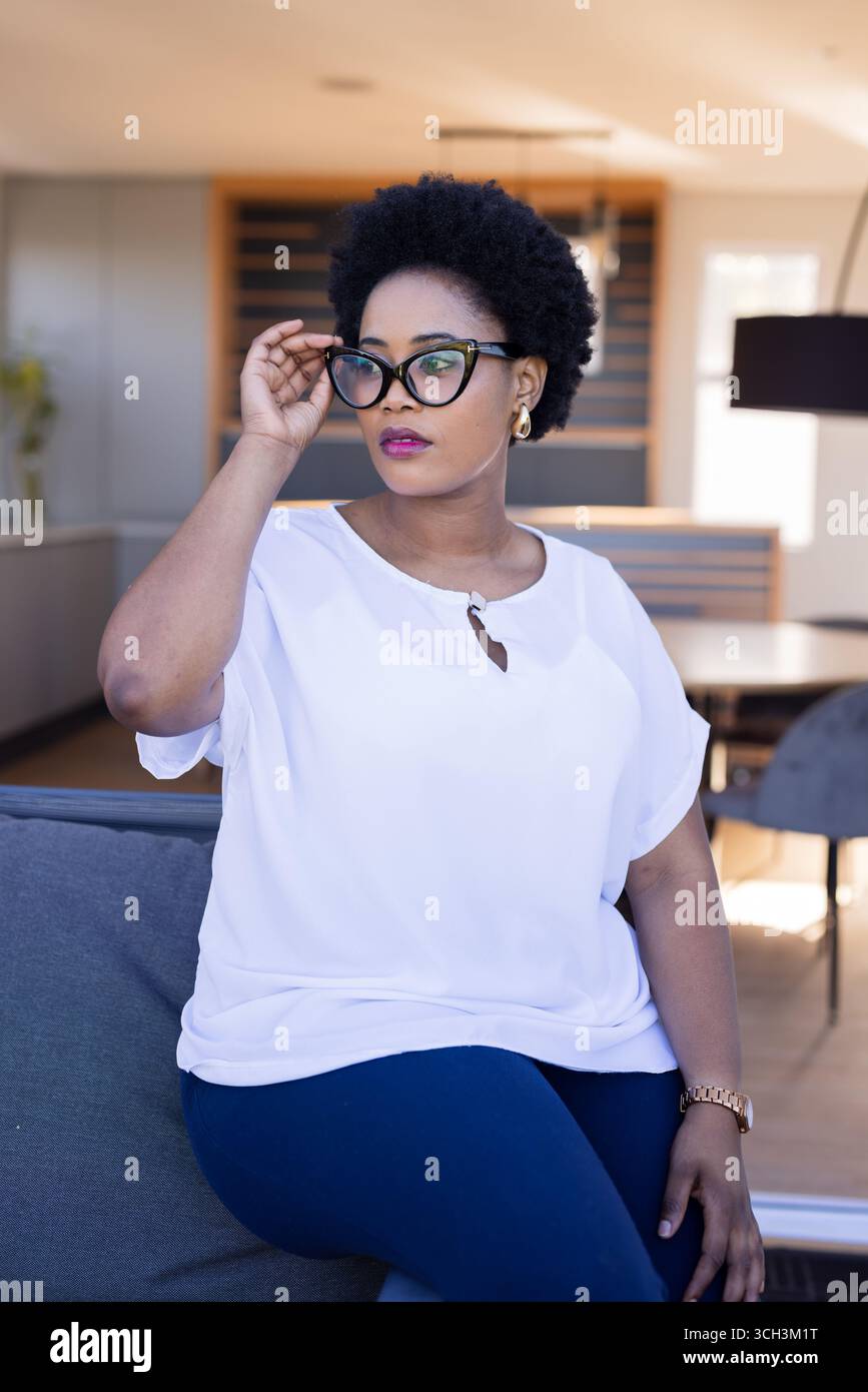 Femme confiante ajustant les lunettes tout en étant assise sur le canapé dans le salon moderne Banque D'Images