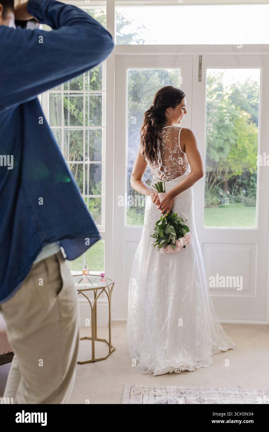 Mariée en robe élégante tenant bouquet, debout par fenêtre, profitant d'un moment serein Banque D'Images