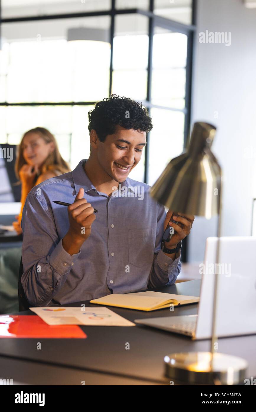 Homme d'affaires souriant à l'aide d'un smartphone et écrivant des notes au bureau Banque D'Images Homme d'affaires souriant à l'aide d'un smartphone et écrivant des notes au bureau Banque D'Images