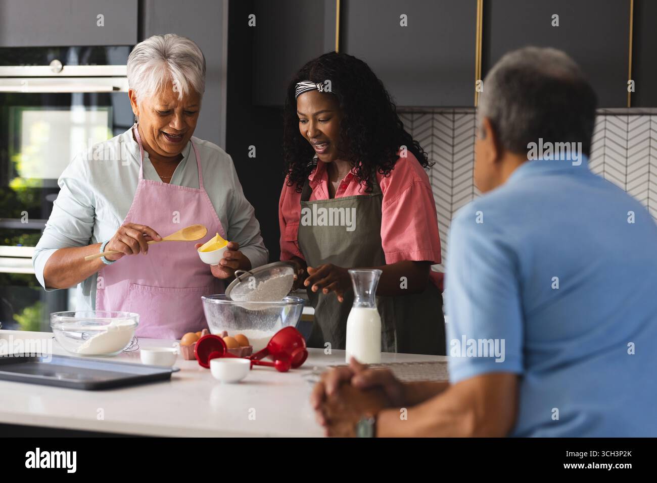 Mère aînée et fille adulte cuisinant dans la cuisine pendant que le père regarde, à la maison Banque D'Images Mère aînée et fille adulte cuisinant dans la cuisine pendant que le père regarde, à la maison Banque D'Images