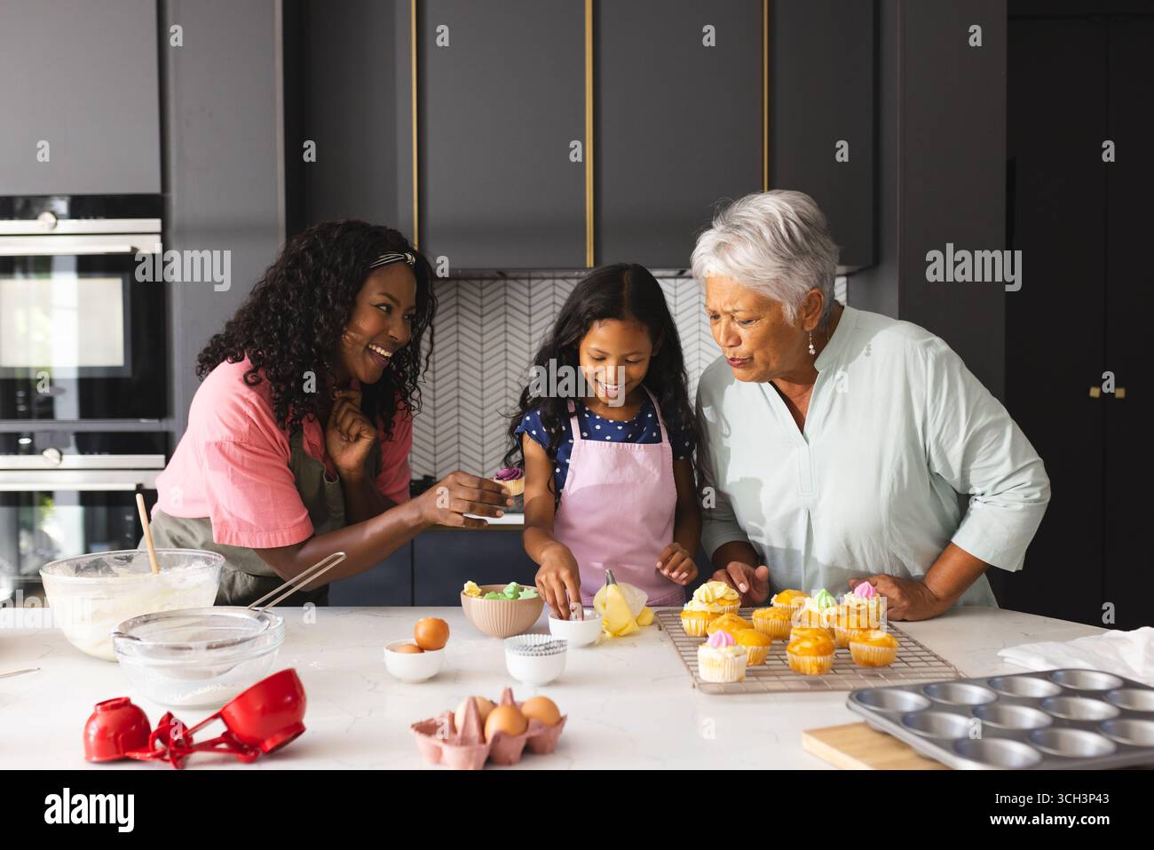 Trois générations heureuses cuisson, souriant et décorant des cupcakes dans une cuisine moderne, à la maison Banque D'Images Trois générations heureuses cuisson, souriant et décorant des cupcakes dans une cuisine moderne, à la maison Banque D'Images