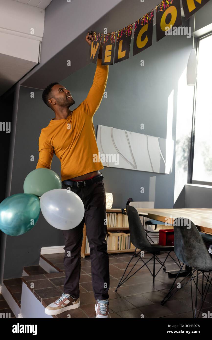 Atteindre l'homme afro-américain accrochant la bannière de bienvenue à l'intérieur du bureau, tenant trois ballons Banque D'Images