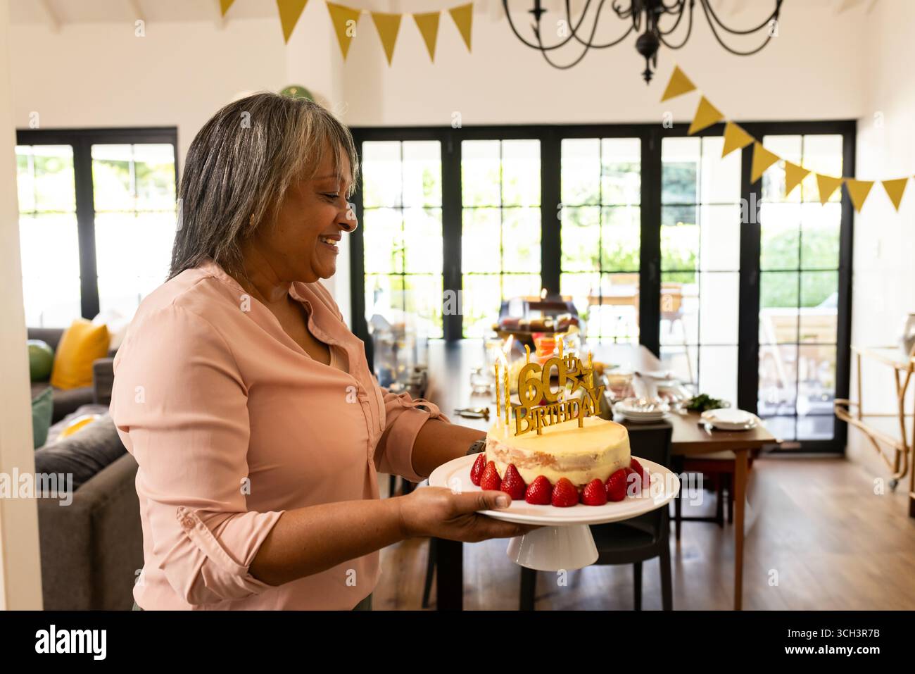 Femme âgée souriante tenant le gâteau du 60ème anniversaire sur stand à la maison, avec des bannières en or Banque D'Images