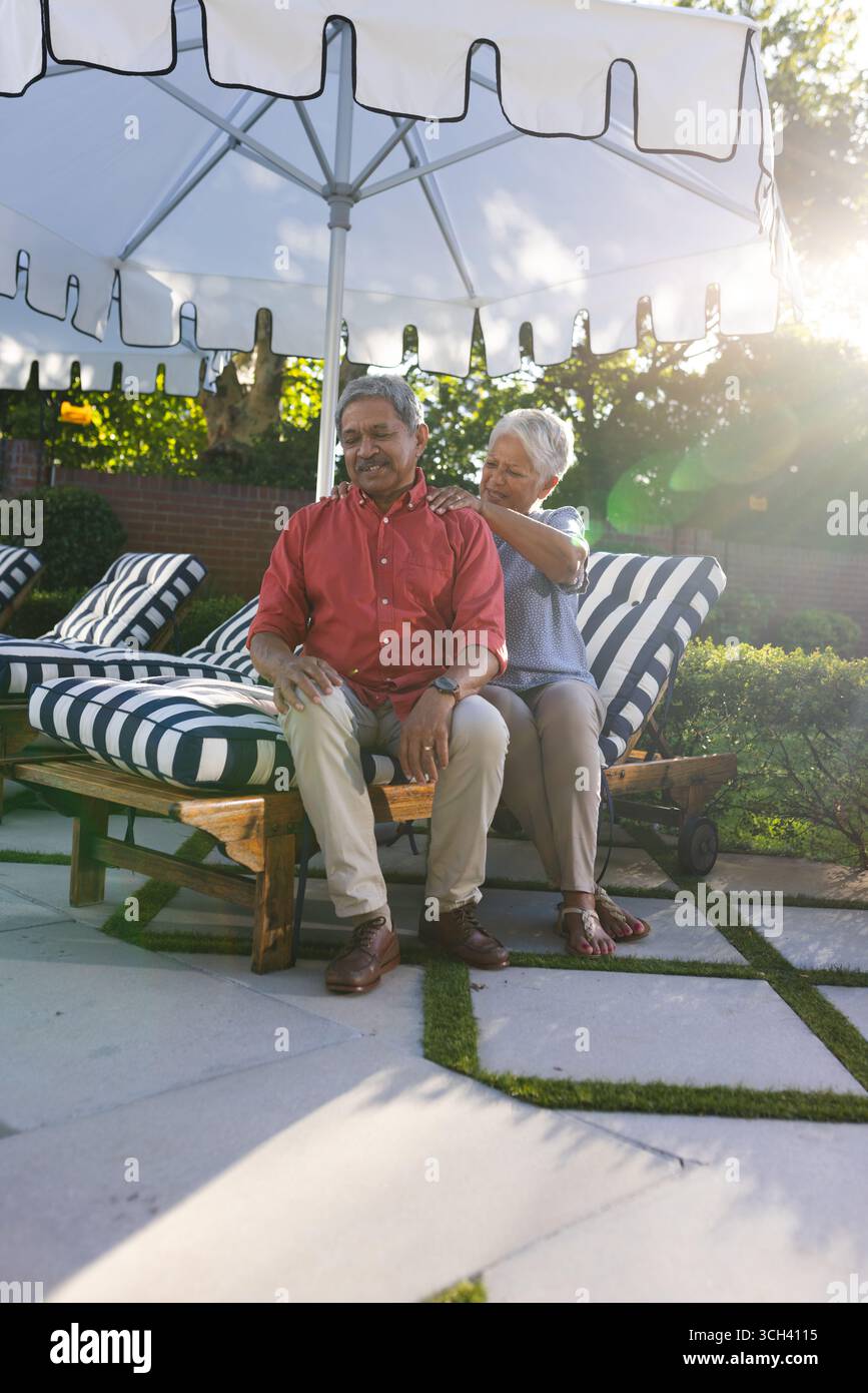 Vieux couple diversifié se relaxant sur des chaises longues sous parasol dans la cour arrière, dans le jardin Banque D'Images