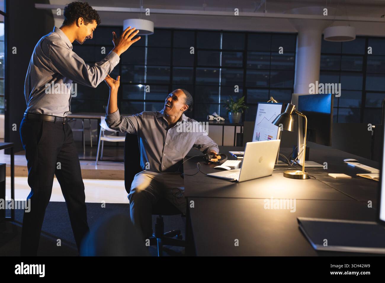 Collègues célébrant le succès avec High-Five dans un bureau moderne la nuit Banque D'Images Collègues célébrant le succès avec High-Five dans un bureau moderne la nuit Banque D'Images