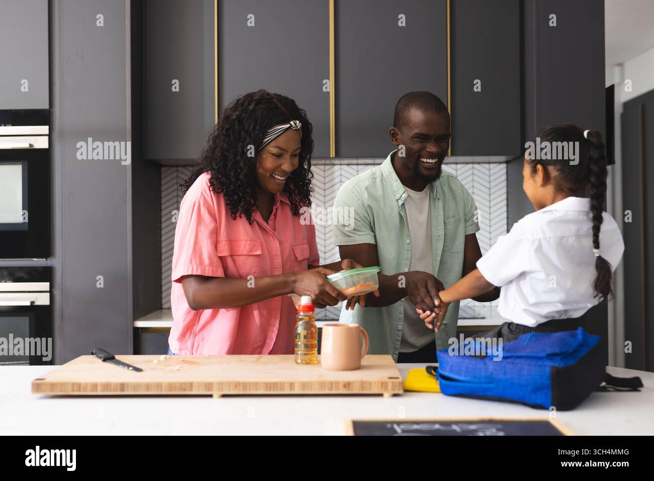 Préparation du déjeuner scolaire dans la cuisine, parents souriant avec leur fille dans la routine du matin, à la maison Banque D'Images Préparation du déjeuner scolaire dans la cuisine, parents souriant avec leur fille dans la routine du matin, à la maison Banque D'Images