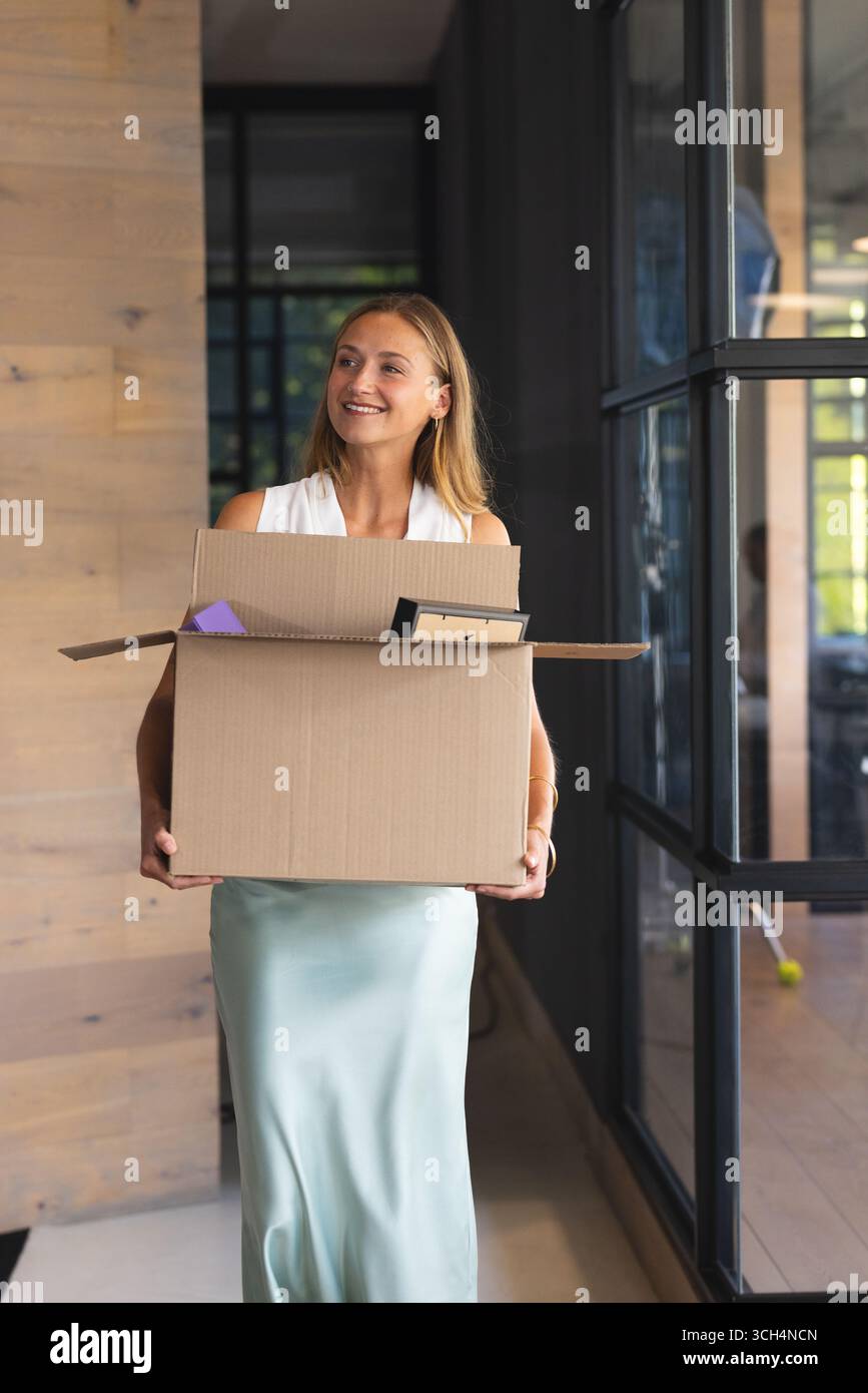 Femme souriante transportant une boîte en carton dans le couloir du bureau, commençant un nouvel emploi Banque D'Images Femme souriante transportant une boîte en carton dans le couloir du bureau, commençant un nouvel emploi Banque D'Images