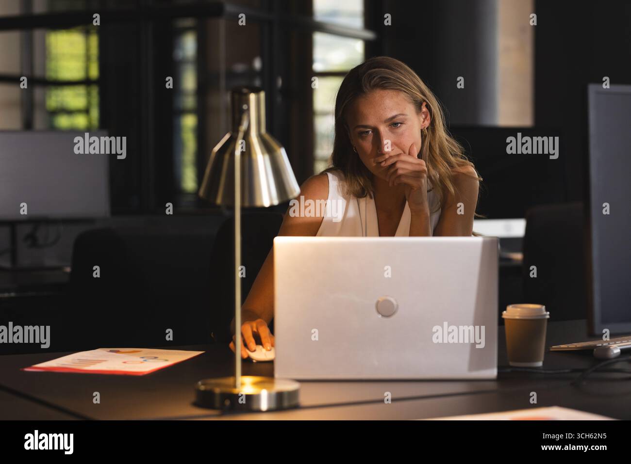 Femme focalisée travaillant tard sur ordinateur portable dans un bureau moderne avec du café Banque D'Images Femme focalisée travaillant tard sur ordinateur portable dans un bureau moderne avec du café Banque D'Images