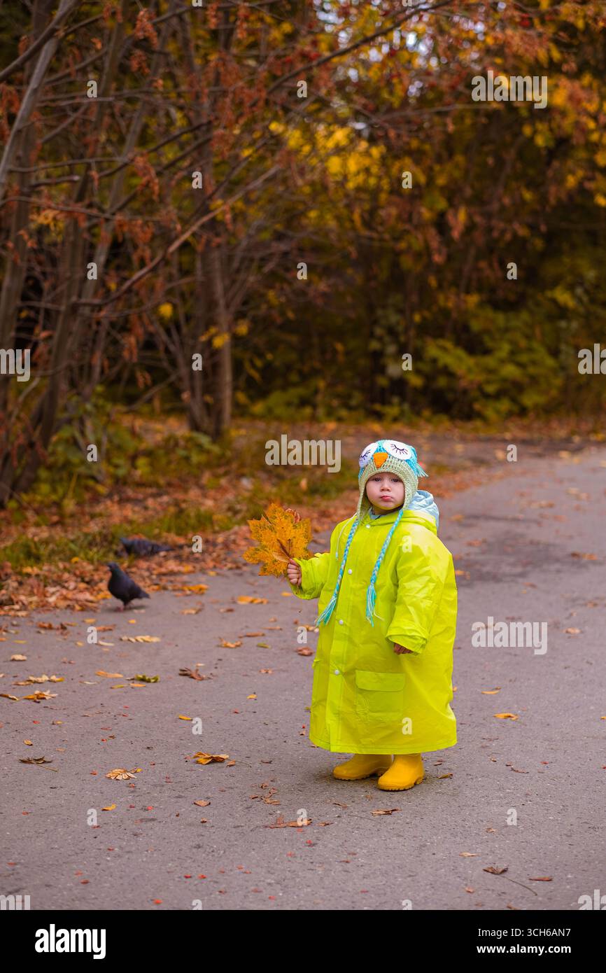 moscou, russie, 06.10.2024 Un enfant dans un imperméable jaune debout sur un chemin tenant une feuille d'automne dans un parc. Concept de saison d'enfance et d'automne. Banque D'Images