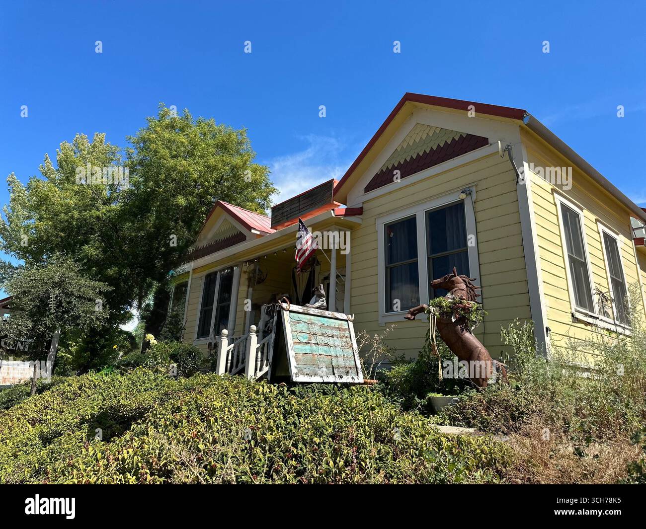 Une charmante maison historique en bois jaune avec garniture de toit rouge, drapeau américain sur le porche, et sculptures de jardin rustique sous un ciel bleu clair. - Image de stock capturée avec un smartphone