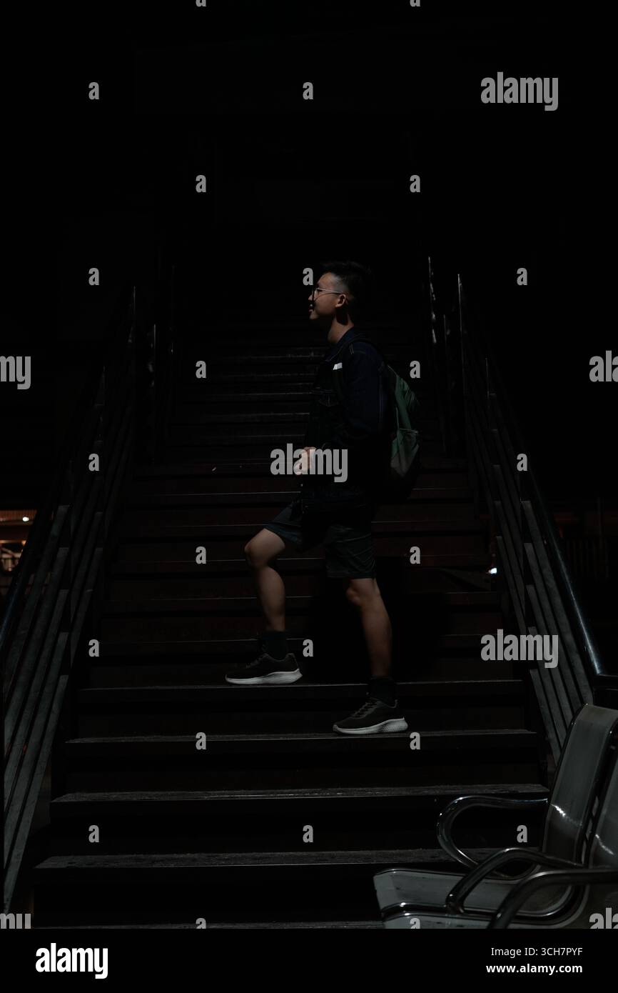Un jeune homme monte un escalier sombre dans un environnement urbain sombre, portant un sac à dos Banque D'Images