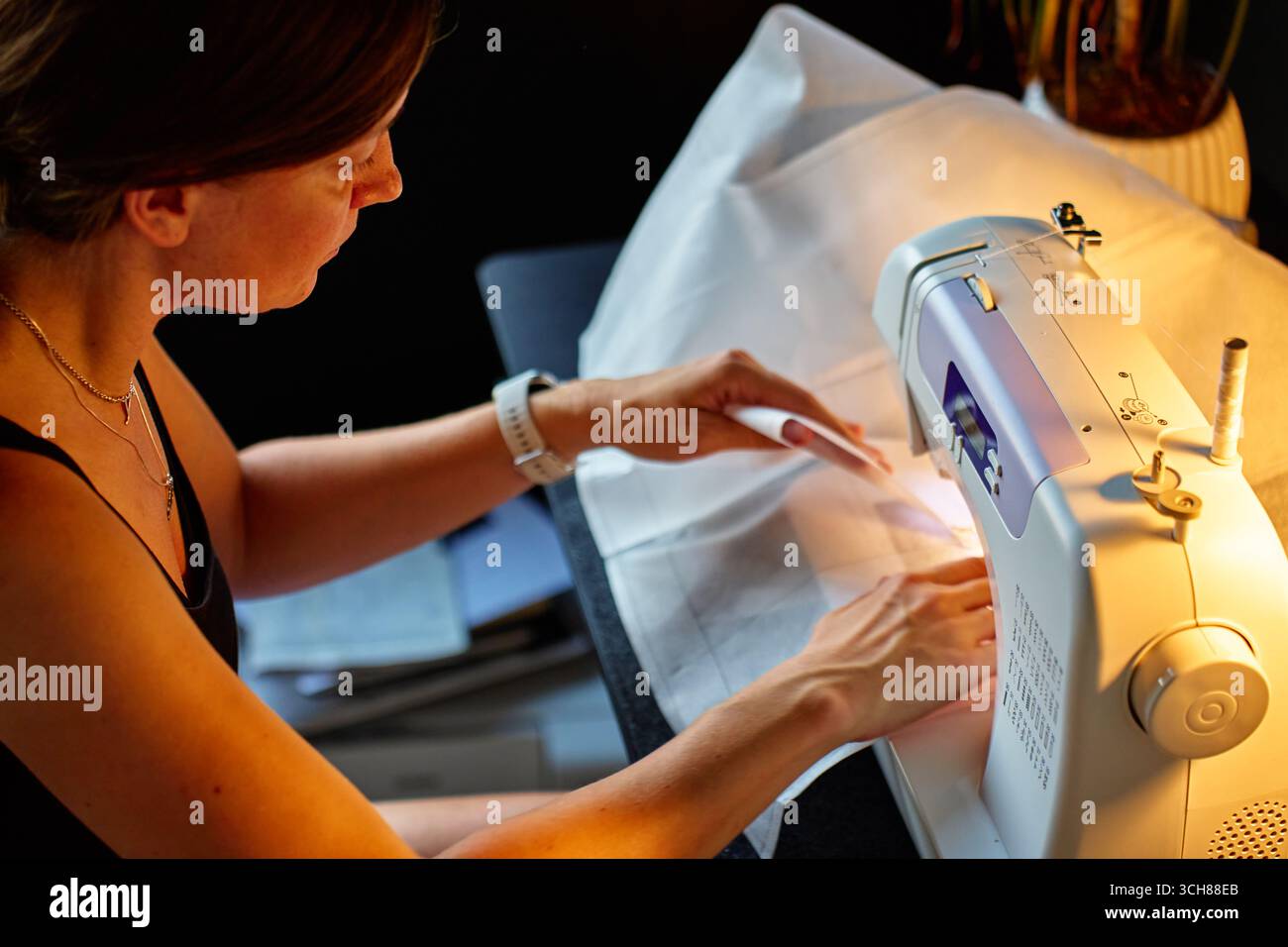 Gros plan de la femme utilisant la machine à coudre moderne pour coudre le tissu à la maison sous la lumière de la lampe. Concept de couture et de couture Banque D'Images