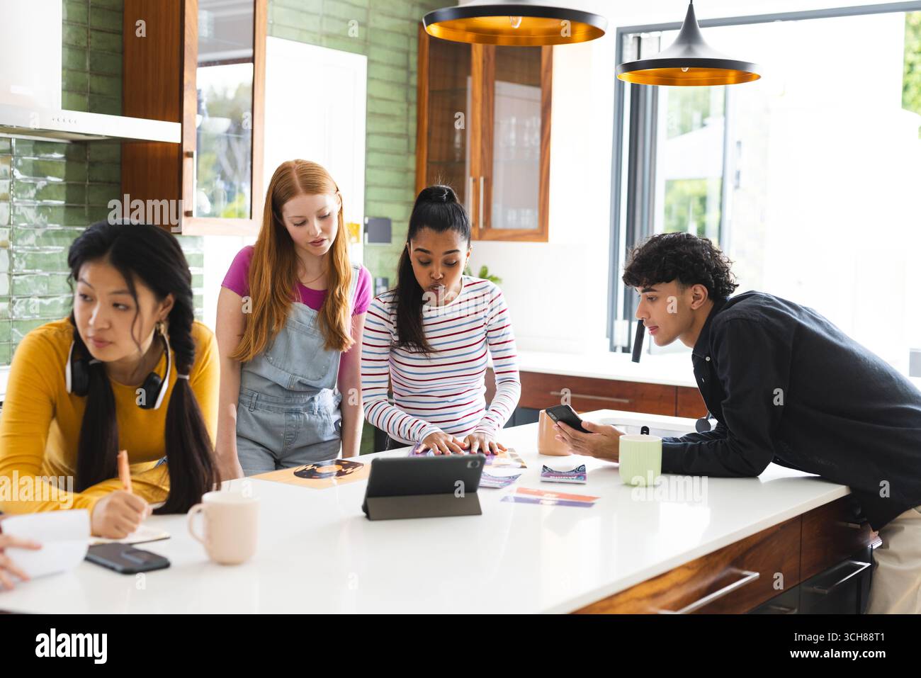 Adolescents collaborant sur un projet scolaire en utilisant une tablette et un smartphone dans une cuisine moderne Banque D'Images