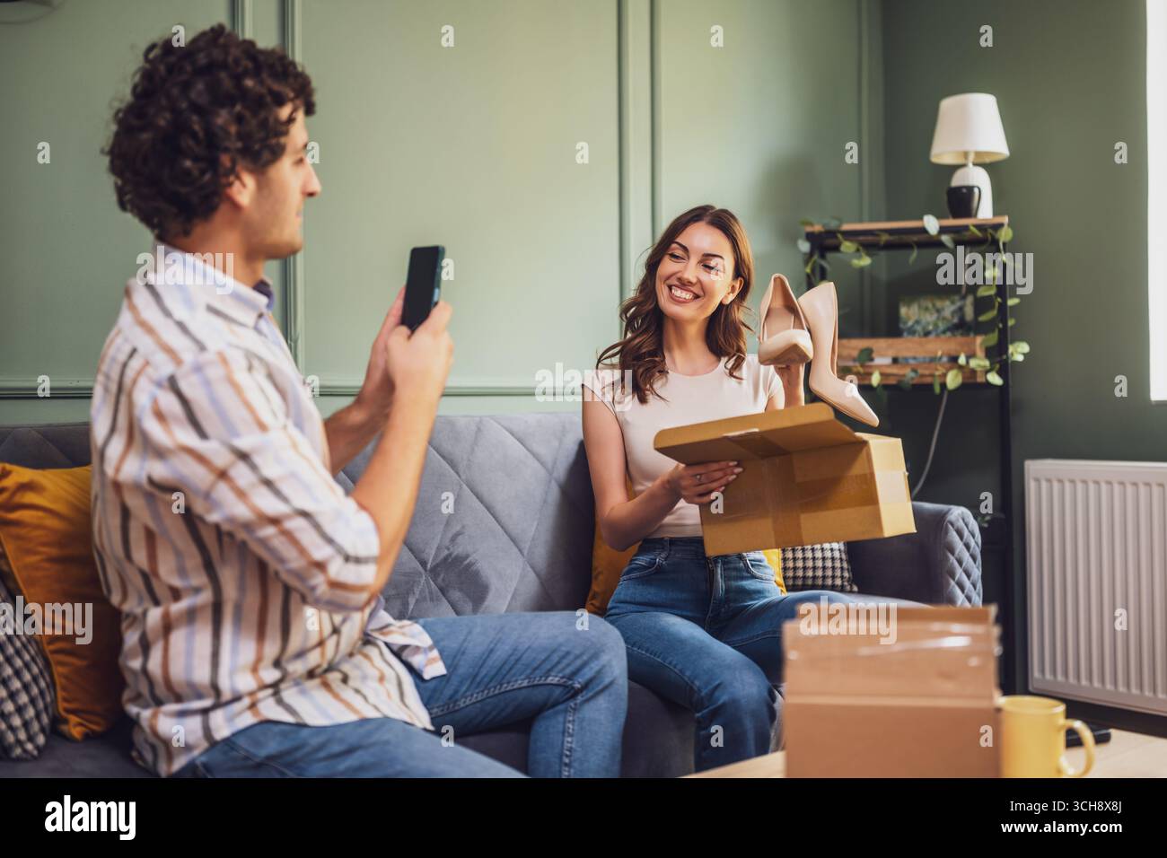 Couple adulte déballage leurs colis livrés à domicile. L'homme prend en photo les nouvelles chaussures de sa femme. Banque D'Images