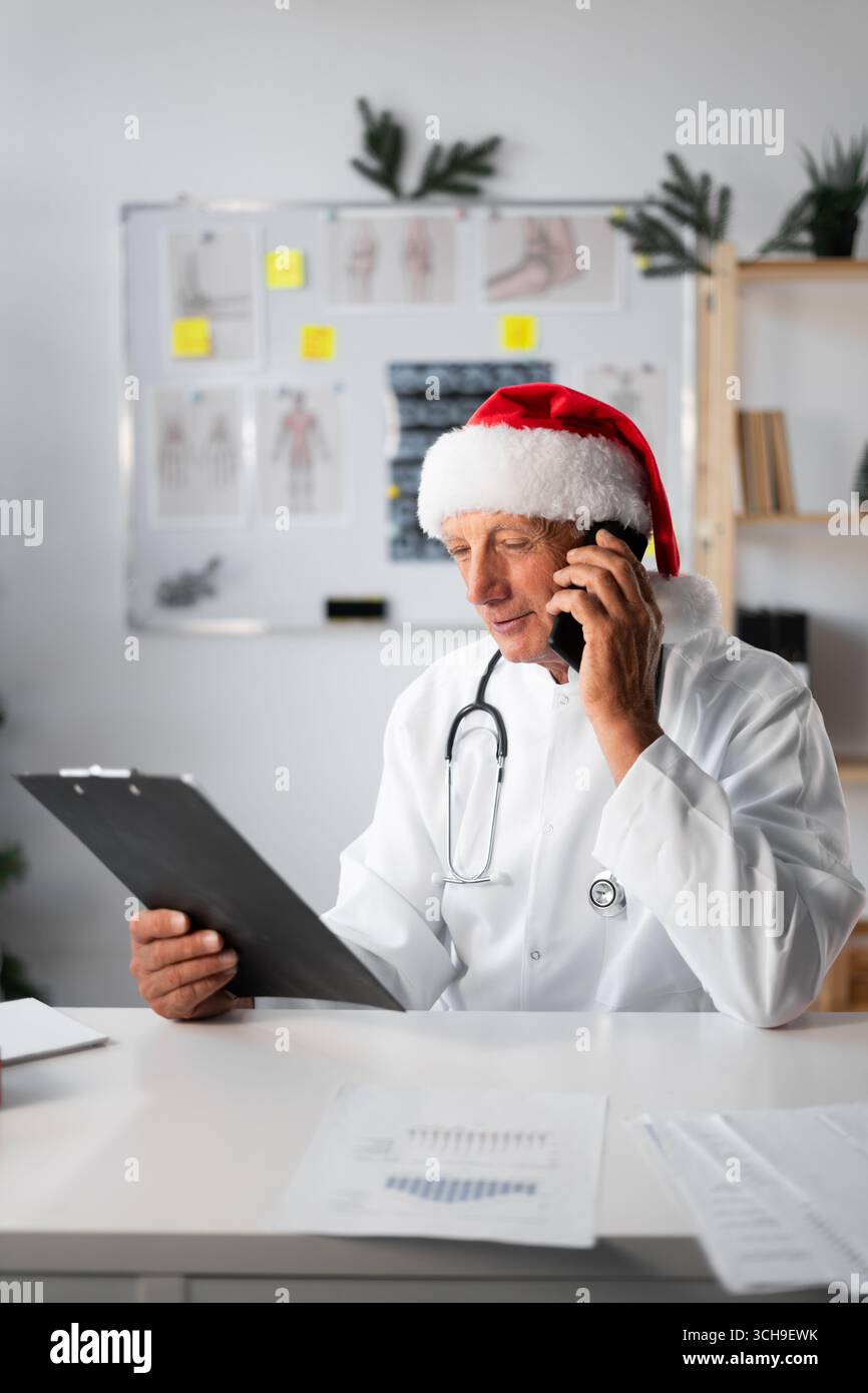 Médecin âgé senior à Santa Hat avec presse-papiers appelant au téléphone à l'hôpital sur le quart de travail de la veille de Noël. Médecine, technologie, Noël et guérison Banque D'Images