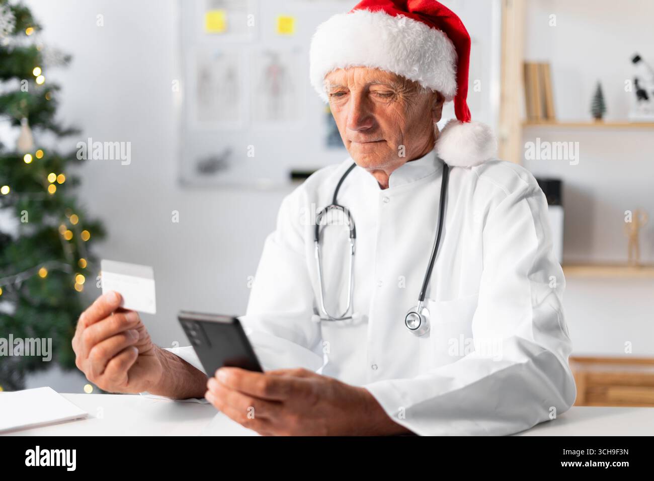 Médecin âgé senior à Santa Hat en utilisant la carte de crédit et le téléphone portable pour les achats en ligne sur le quart de travail à l'hôpital la veille de Noël. médecine, hol Banque D'Images