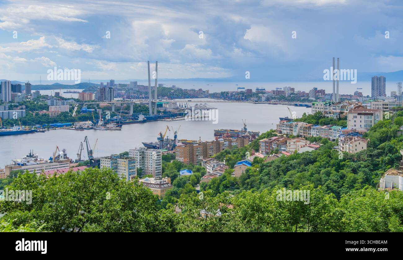 Vue panoramique de Vladivostok, Russie, avec le Pont d'Or sur la baie de la Corne d'Or, les grues portuaires, les navires et le paysage urbain à flanc de colline. Banque D'Images
