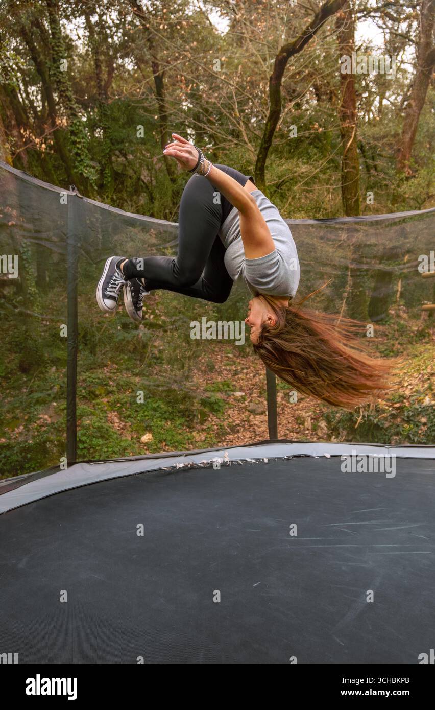 Jeune femme vêtue de vêtements de sport faisant un backflip sur un trampoline dans une forêt en plein air dans les montagnes, profitant du moment. Banque D'Images