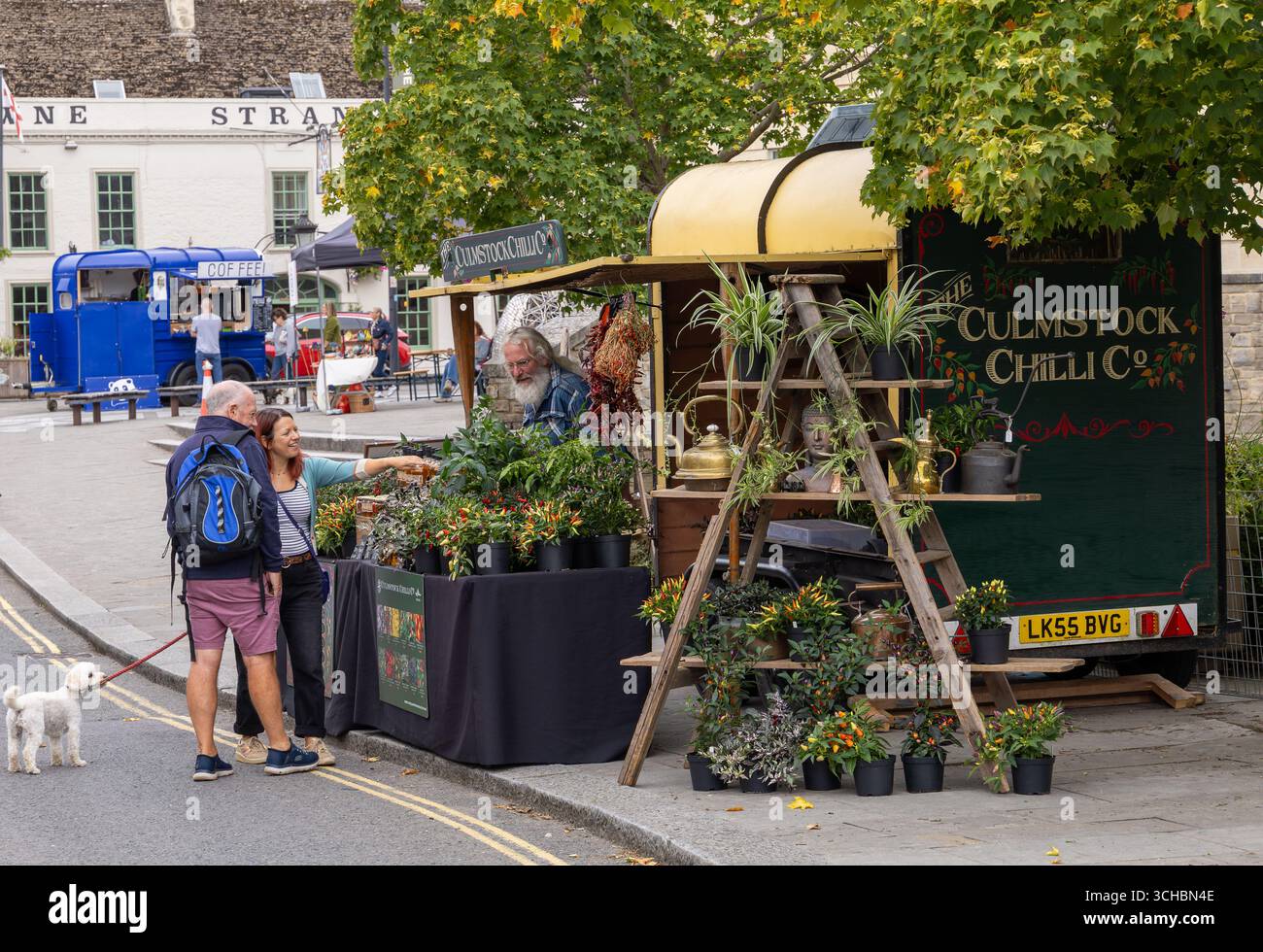 Un couple avec un petit chien magasins pour les plantes de piment à un étal de marché en plein air avec une remorque vintage et diverses plantes en pot affichées Banque D'Images