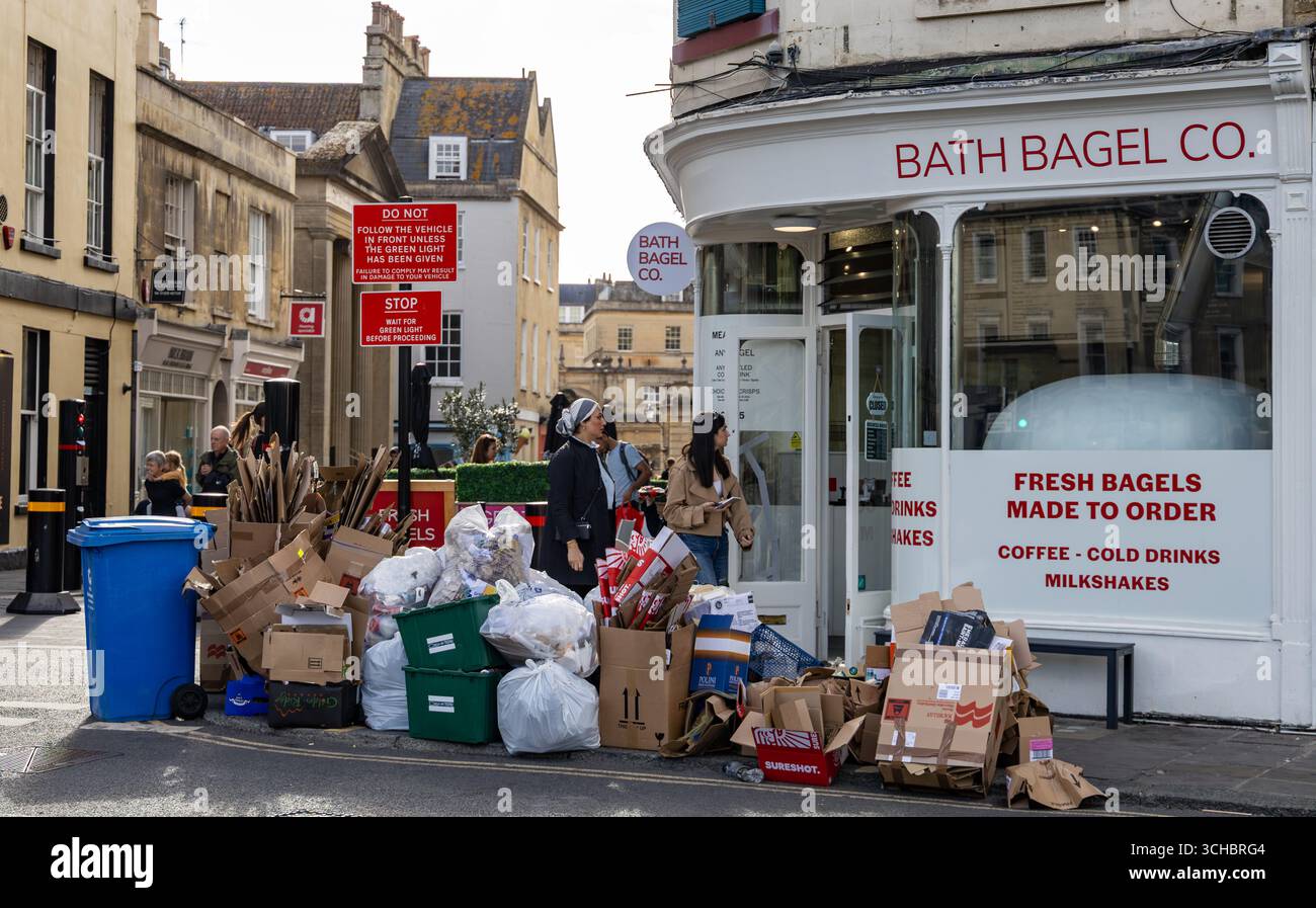 Un coin de rue à Bath, en Angleterre, avec des sacs à ordures / poubelles et du carton empilés dehors prêts à être ramassés dans un magasin de bagels avec des gens qui passent Banque D'Images