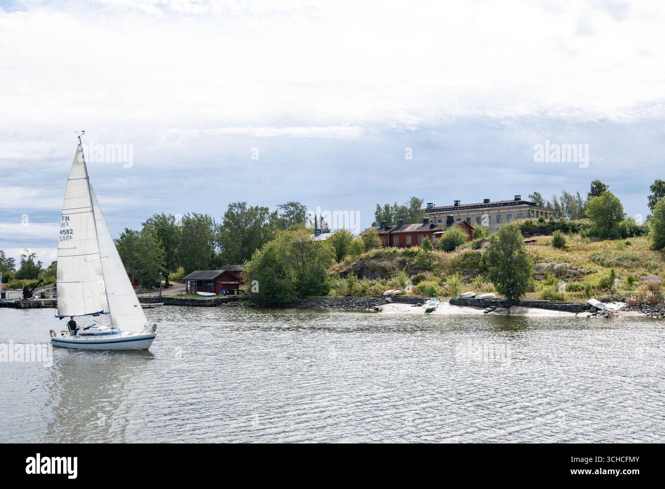 Voilier naviguant dans le port d'Helsinki avec une île rocheuse et des bâtiments finlandais traditionnels en arrière-plan Banque D'Images