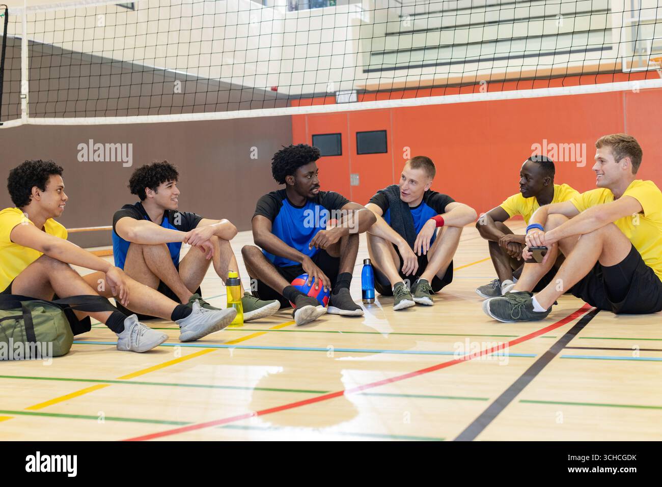 Divers coéquipiers masculins assis sur le terrain de gymnastique sous le filet avec volley-ball, bouteilles d'eau et sac de sport. Athlètes, travail d'équipe, camaraderie, entraînement, fitnes Banque D'Images Divers coéquipiers masculins assis sur le terrain de gymnastique sous le filet avec volley-ball, bouteilles d'eau et sac de sport. Athlètes, travail d'équipe, camaraderie, entraînement, fitnes Banque D'Images