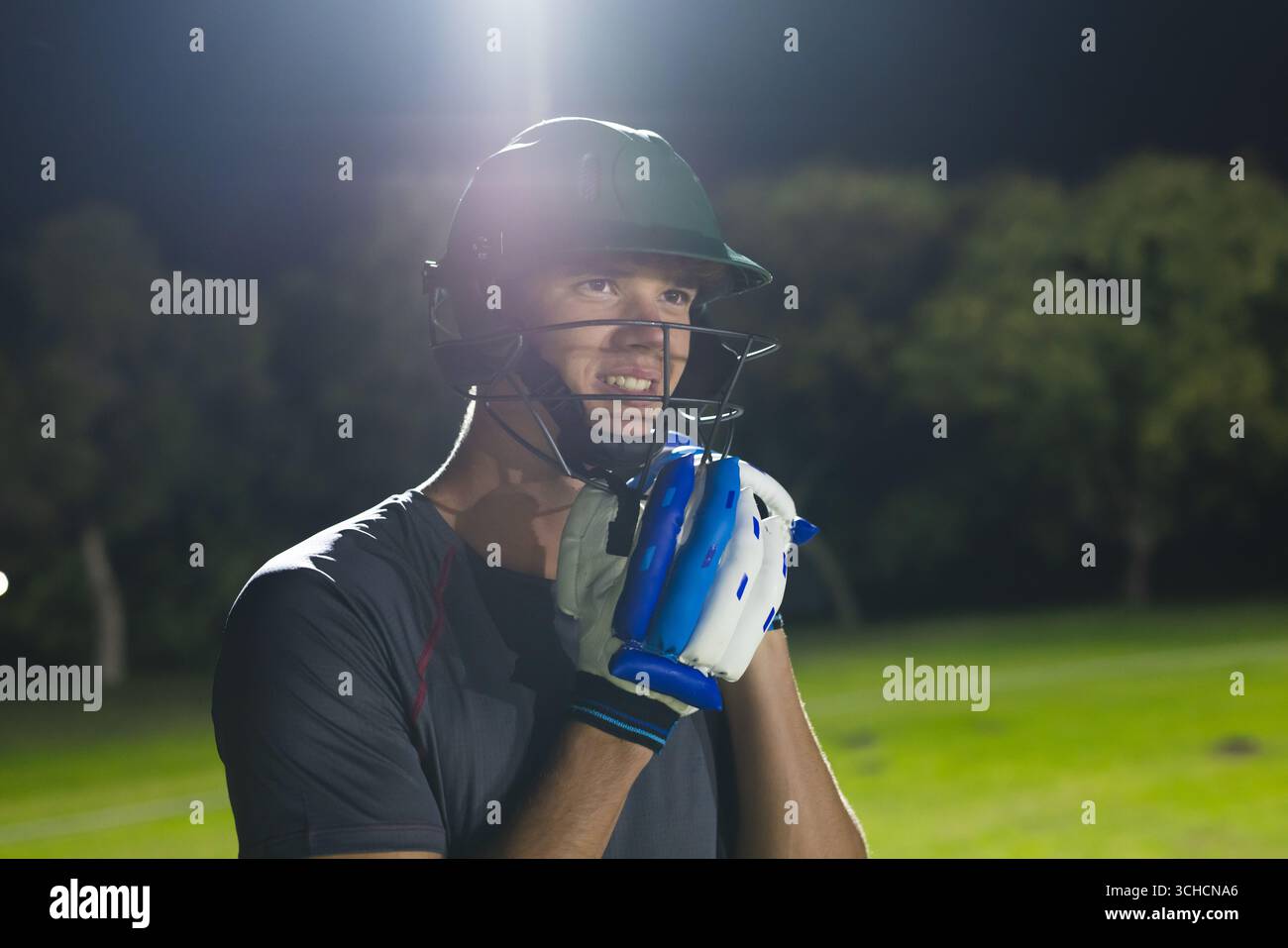 Athlète masculin debout sur le terrain la nuit sous des projecteurs portant un casque de cricket et des gants de frappe. Athlète, sports, extérieur, nuit, stade, compet Banque D'Images