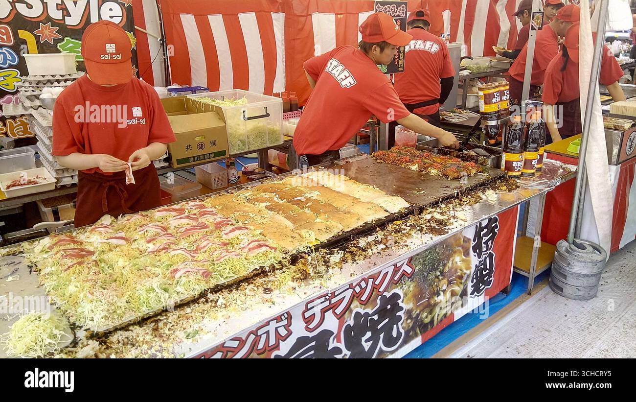 Le personnel d'un stand de nourriture de festival japonais animé préparant et cuisinant Okonomiyaki sur une grande plaque chauffante. Banque D'Images