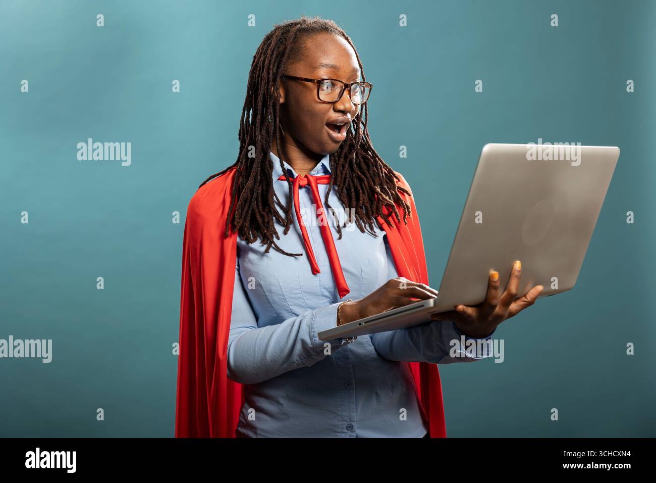Une femme afro-américaine choquée dans une cape rouge regarde son ordinateur portable avec de larges yeux. Modèle féminin noir avec réaction expressive véhiculant de l'excitation et des nouvelles positives inattendues. Banque D'Images