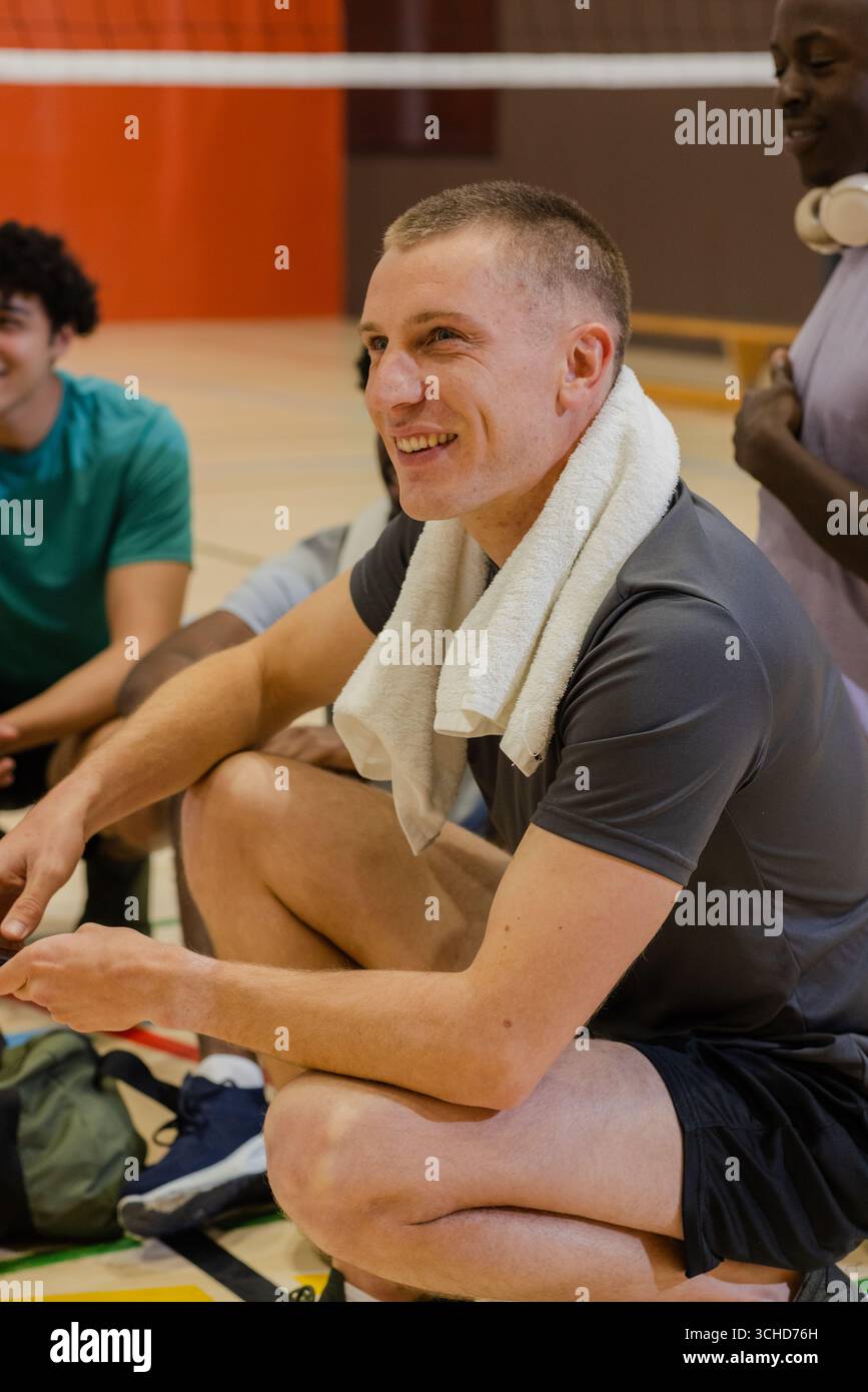 Divers coéquipiers masculins accroupis sur le terrain avec une serviette drapée autour du cou et un sac vert près du filet. Athlétique, camaraderie, travail d'équipe, sportsgym, récupération, Banque D'Images Divers coéquipiers masculins accroupis sur le terrain avec une serviette drapée autour du cou et un sac vert près du filet. Athlétique, camaraderie, travail d'équipe, sportsgym, récupération, Banque D'Images