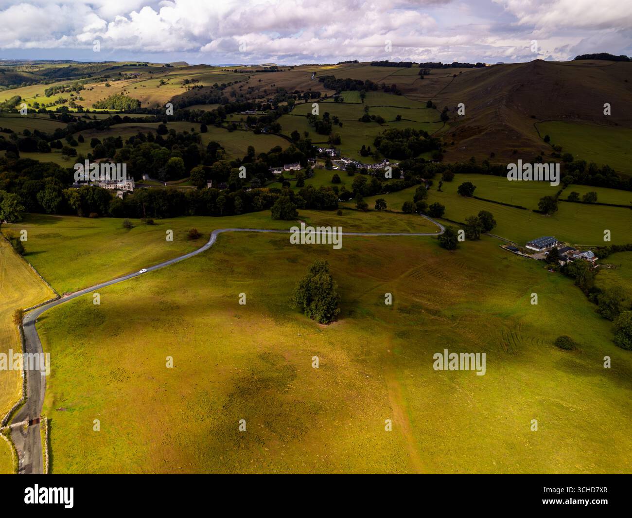 Vue aérienne d'une voiture blanche conduisant sur une route sinueuse à travers des collines ondulantes et des champs verdoyants dans le Peak District, avec des nuages spectaculaires au-dessus Banque D'Images