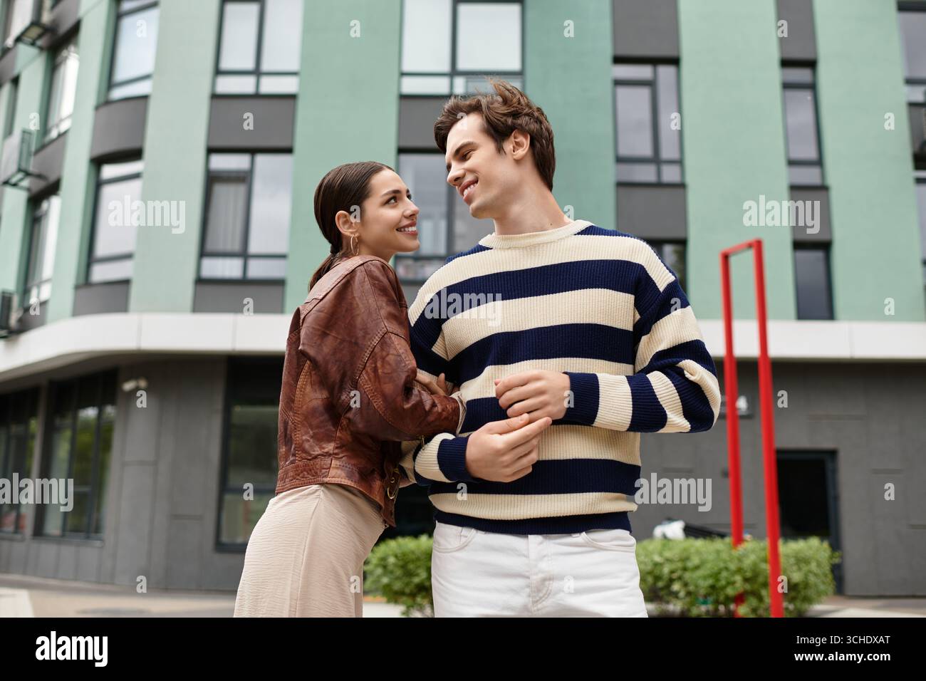 Un jeune couple se sourit, partageant une étreinte chaleureuse dans un endroit urbain dynamique. Banque D'Images