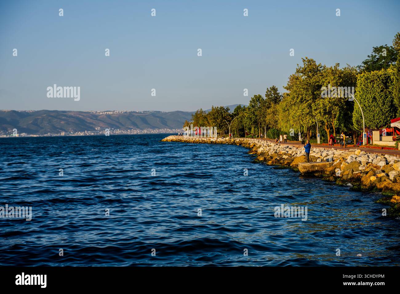 Les gens marchent et se détendent le long de la promenade balnéaire bordée d'arbres à Karamürsel, Kocaeli, Turquie, avec des montagnes visibles à travers le golfe de İzmit. Ka Banque D'Images