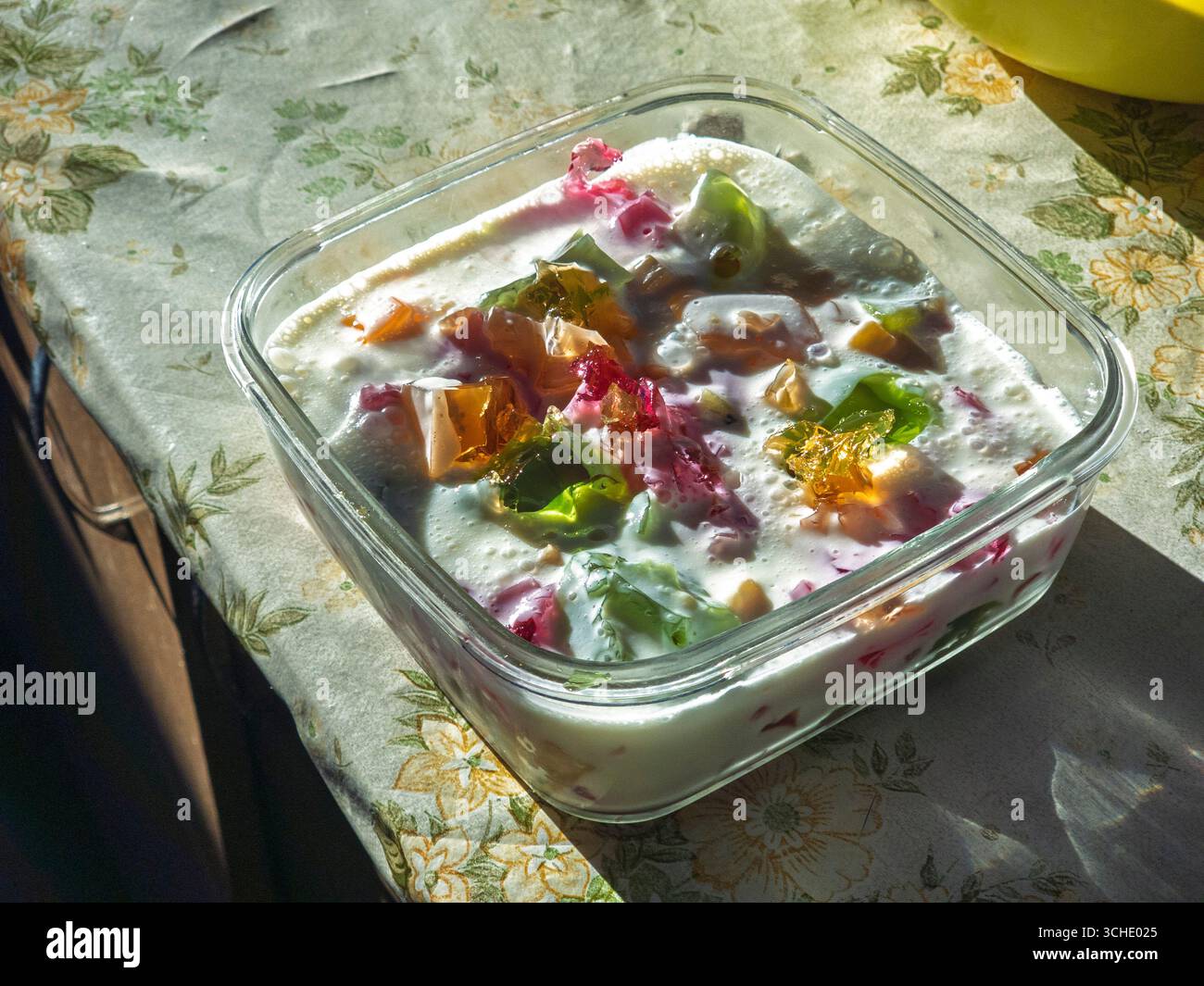Dessert rafraîchissant et coloré en gélatine mélangé avec du yaourt est présenté dans un récipient en verre carré, assis sur une table avec une nappe florale, creat Banque D'Images Dessert rafraîchissant et coloré en gélatine mélangé avec du yaourt est présenté dans un récipient en verre carré, assis sur une table avec une nappe florale, creat Banque D'Images