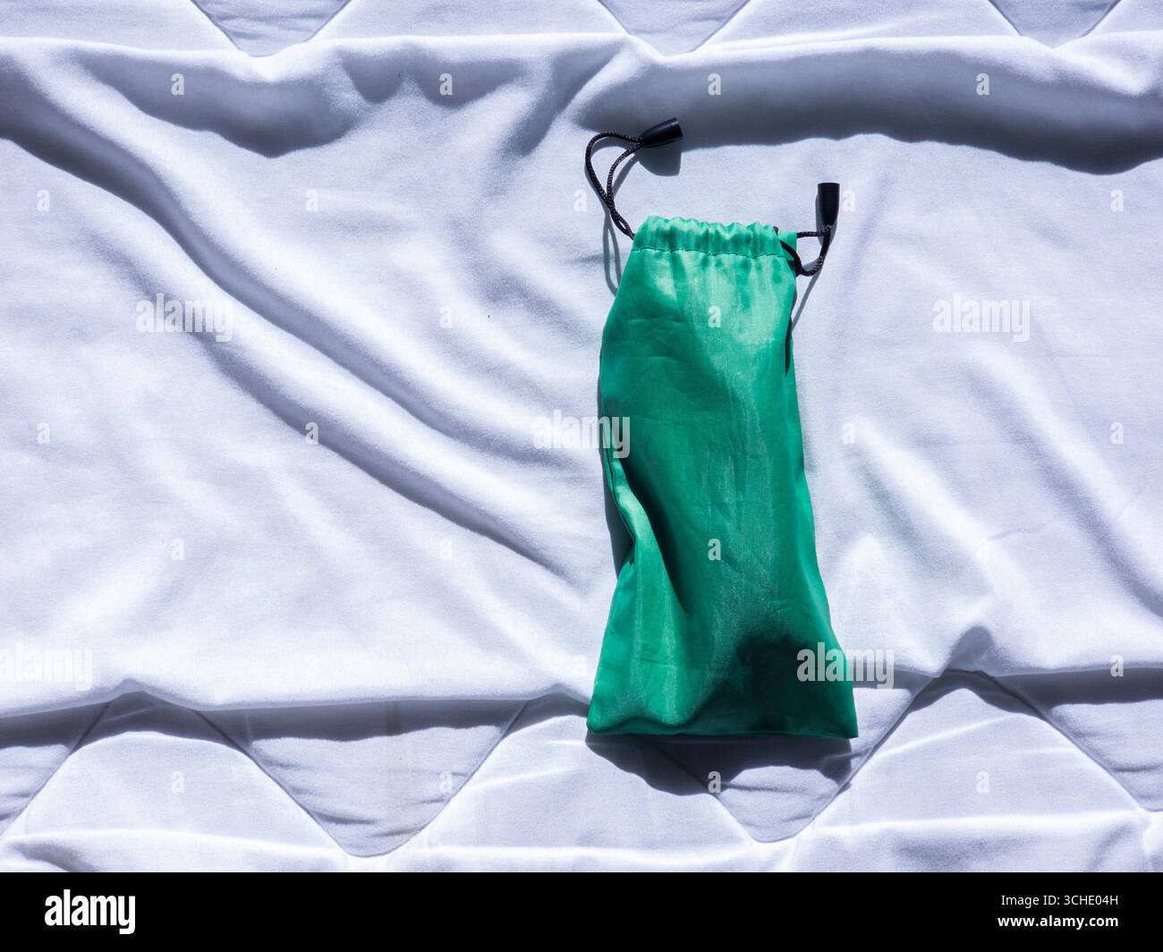 Petit sac à cordon vert reposant sur des feuilles blanches, se prélasser au soleil brillant, projetant des ombres fortes et des reflets qui améliorent la texture lisse Banque D'Images