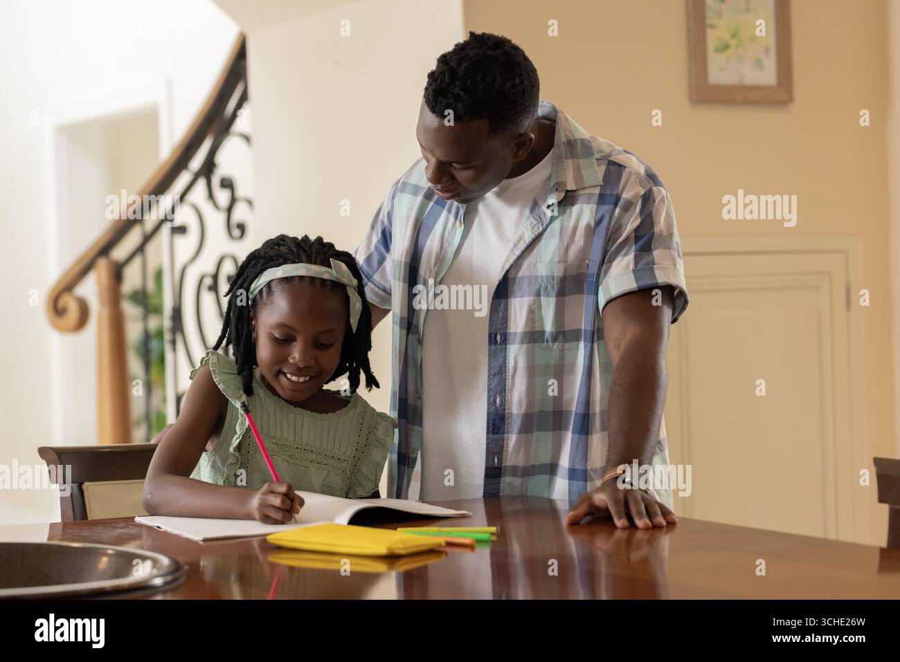 Père afro-américain aidant sa fille aux devoirs à la maison, les deux souriant chaleureusement Banque D'Images Père afro-américain aidant sa fille aux devoirs à la maison, les deux souriant chaleureusement Banque D'Images