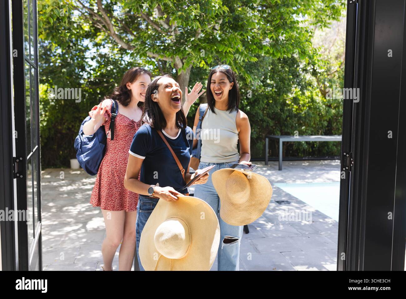 Trois amies féminines diverses riant et entrant dans la maison avec des chapeaux de soleil et des sacs à dos Banque D'Images
