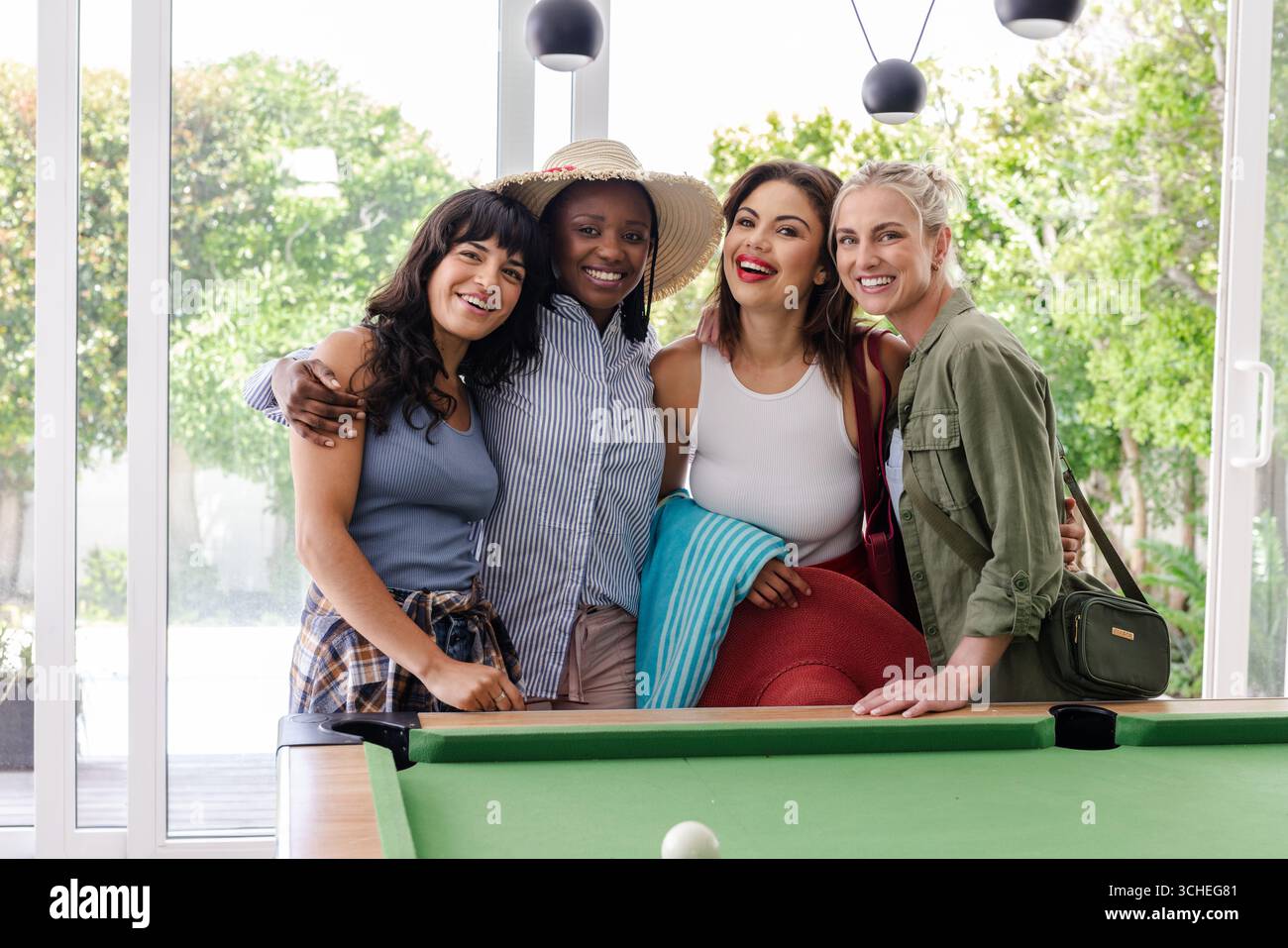 Groupe diversifié d'amies féminines diverses souriant et embrassant près de la table de billard à la maison Banque D'Images Groupe diversifié d'amies féminines diverses souriant et embrassant près de la table de billard à la maison Banque D'Images