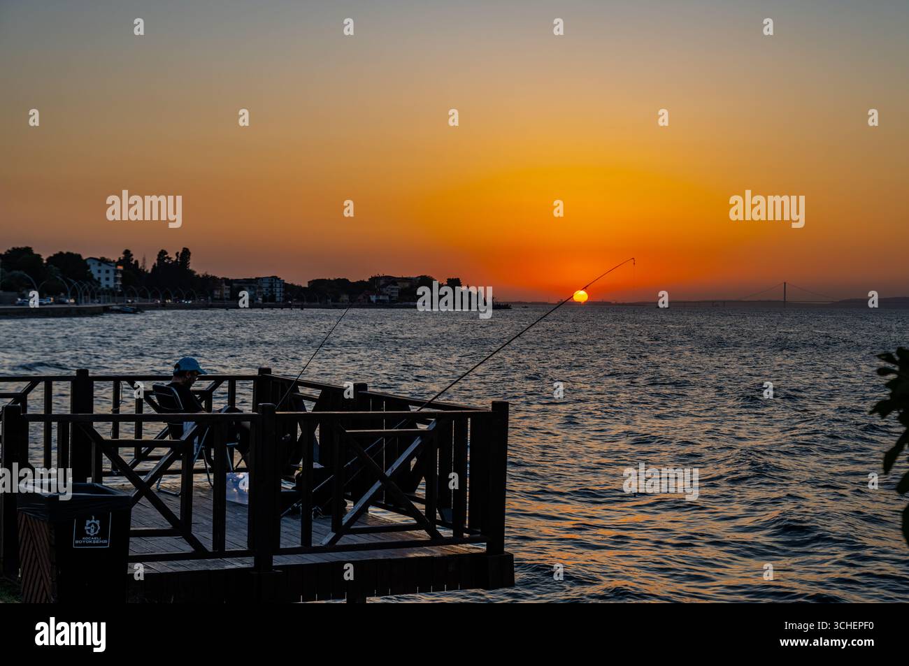Les pêcheurs sont assis sur un pont en bois avec des cannes à pêche pendant le coucher du soleil sur le golfe de İzmit, avec le pont Osman Gazi visible au loin de Karamür Banque D'Images