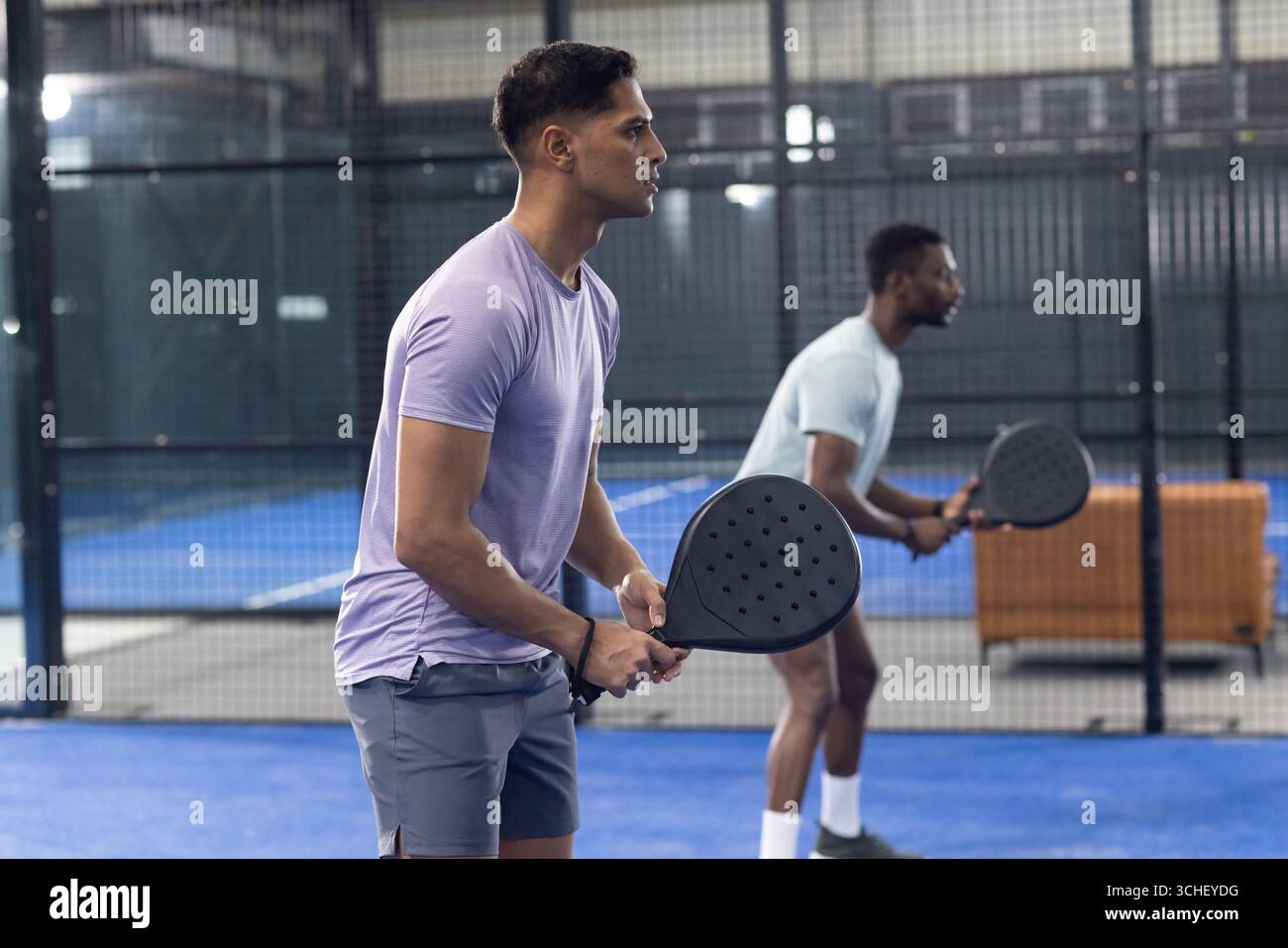 Divers amis masculins saisissant des raquettes de padel, regardant la balle entrante sur le terrain bleu avec banc Banque D'Images